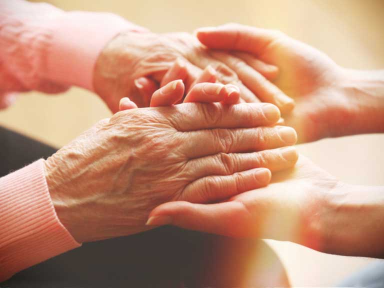 Close-up of a younger person gently holding the hands of an older person, symbolizing care, support, and compassion. Warm lighting creates a comforting atmosphere.