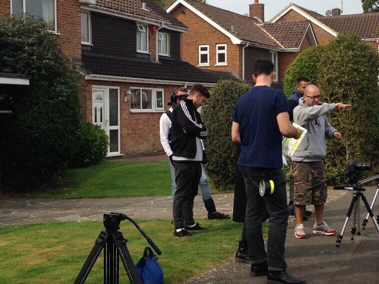 A small film crew stands on a suburban sidewalk near a camera and tripod, with houses and green bushes in the background. One person points while others look on or check equipment.