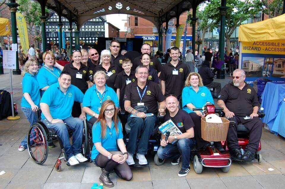 A group of people, some in wheelchairs, pose and smile together outdoors under a decorative gazebo. Some wear matching blue shirts, others wear black shirts, with event tents and banners visible in the background.