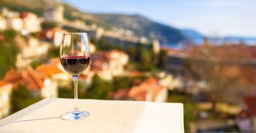 A glass of red wine sits on a white ledge, with a blurred background of sunlit buildings and a scenic hillside, suggesting a peaceful outdoor setting.