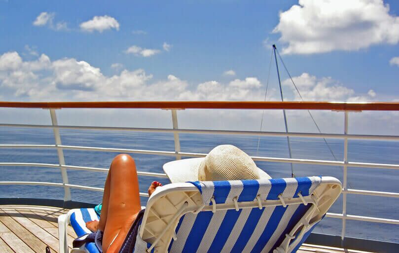 A person in a sunhat relaxes on a blue and white striped lounge chair on a ship deck, facing the ocean under a bright blue sky with scattered clouds. A sailboat mast is visible in the distance.
