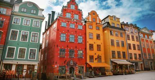 Colorful historic buildings with ornate facades stand side by side in a European city square under a blue sky with scattered clouds; outdoor seating is visible in front of some buildings.