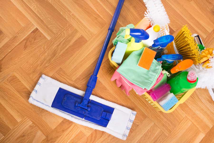 A blue mop lies on a wooden floor next to a yellow basket filled with cleaning supplies, including spray bottles, sponges, cloths, gloves, and brushes.