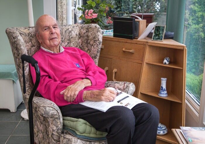 An elderly man sits in a floral armchair by a window, wearing a bright pink sweater. He holds a magazine on his lap, with a cane resting beside him. A wooden cabinet and potted plant are in the background.