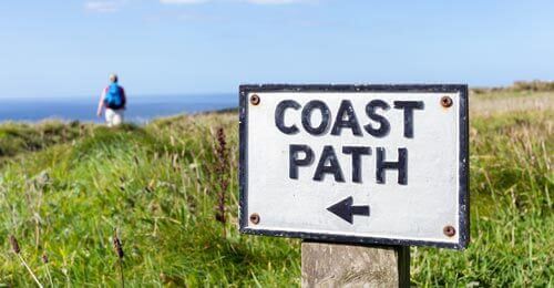 A close-up of a Coast Path sign with an arrow pointing left, set in grassy countryside. In the background, a person with a backpack walks along a trail toward the distant sea.