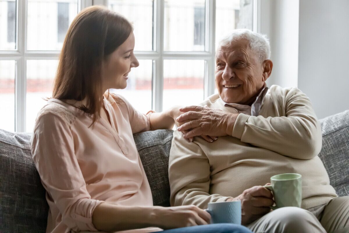 A young woman and an older man sit together on a couch, smiling and having a conversation while holding mugs. Sunlight streams through a window behind them, creating a warm and relaxed atmosphere.