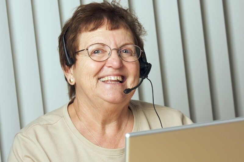 An older woman wearing glasses and a headset smiles while sitting in front of a laptop, with vertical blinds in the background.