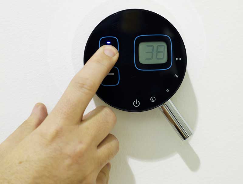 A person adjusts a modern black digital thermostat mounted on a white wall, displaying the number 38 on its screen.
