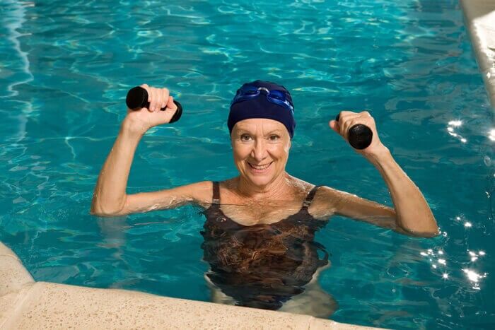 An older woman in a swimsuit and swim cap is standing in a pool, smiling and holding water dumbbells above her shoulders, appearing to exercise in the water.