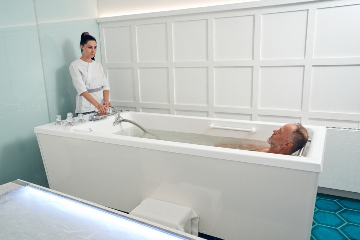 A woman in a white uniform stands by a large white bathtub, adjusting the tap, while a man relaxes in the bath in a brightly lit, modern room.