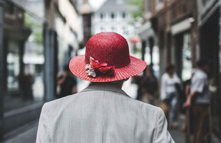 A person wearing a red hat with a flower decoration and a striped jacket is walking down a city street, seen from behind. The background is blurred, with people and buildings visible.