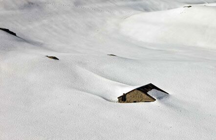 A small house almost completely buried in deep snow, with only its roof and chimney visible above the surrounding white landscape.