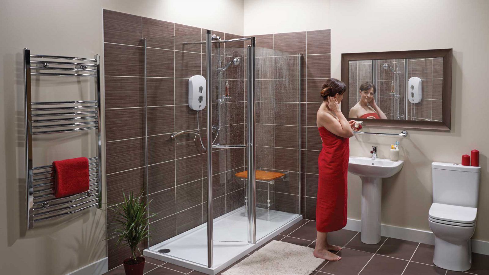 A woman in a red towel stands at a sink in a modern bathroom with a glass shower, wall-mounted electric shower unit, toilet, towel rack, potted plant, and a large mirror above the sink.