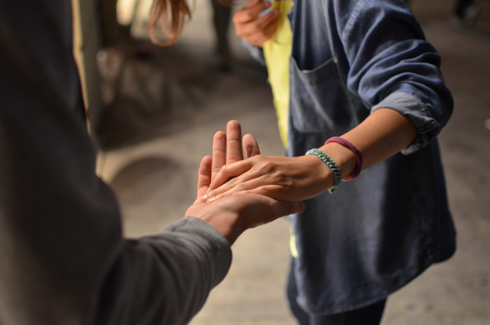 Two people holding hands gently, one reaching out to grasp the others palm. Both wear casual clothing, and only their arms and torsos are visible. The background is blurred.