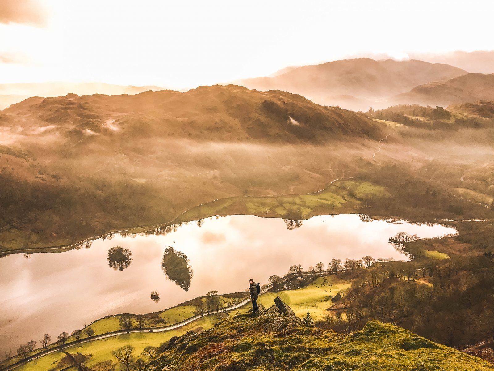 A lone hiker stands on a grassy hilltop overlooking a reflective lake surrounded by rolling hills and mist, with soft golden light illuminating the landscape.