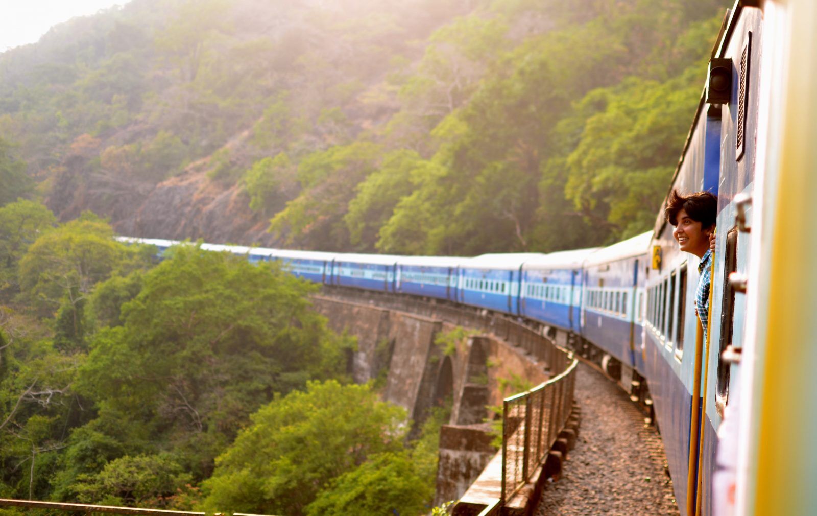 A blue train winds through lush green hills, crossing a bridge. A smiling person leans out of a window, enjoying the scenic view. The landscape is sunlit and vibrant.