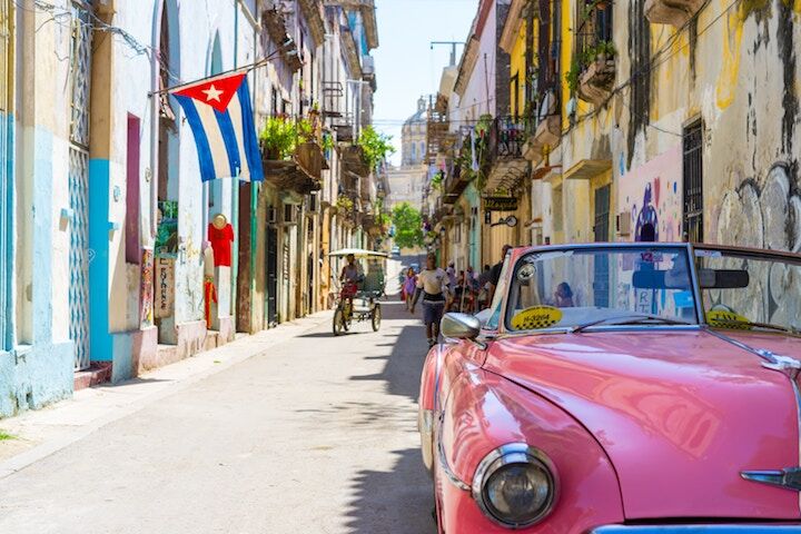 A vibrant street in Cuba with a pink vintage convertible car parked, colorful old buildings, a Cuban flag hanging, and people walking and riding bicycles under bright sunlight.