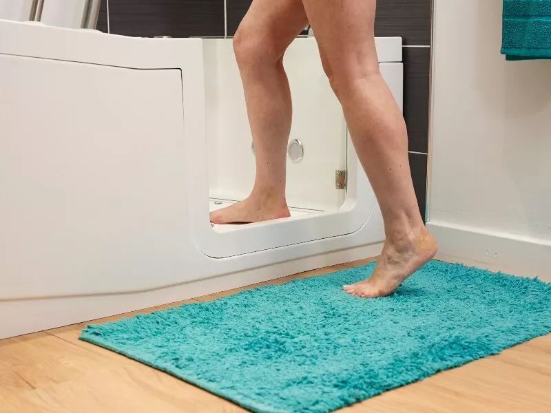 A person steps into a walk-in bathtub, with one foot inside and one foot outside on a turquoise bath mat, in a bathroom with wood flooring and tiled walls.