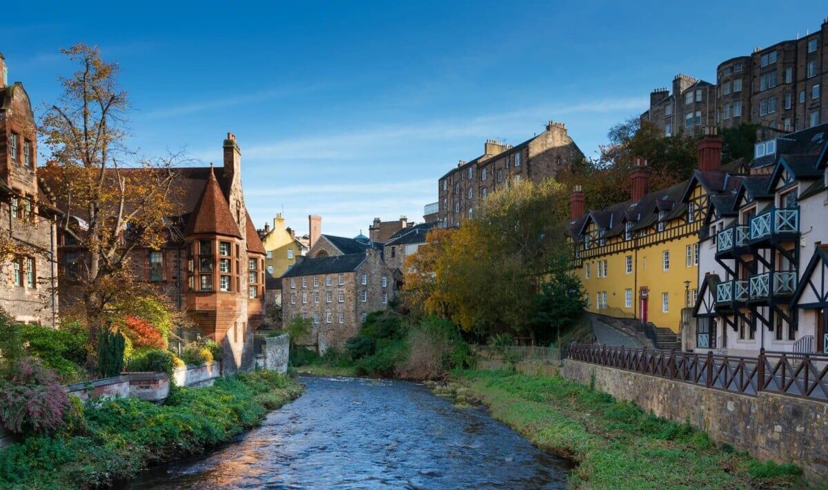 A narrow river flows through a picturesque village lined with historic, colorful buildings and lush greenery, beneath a bright blue sky.