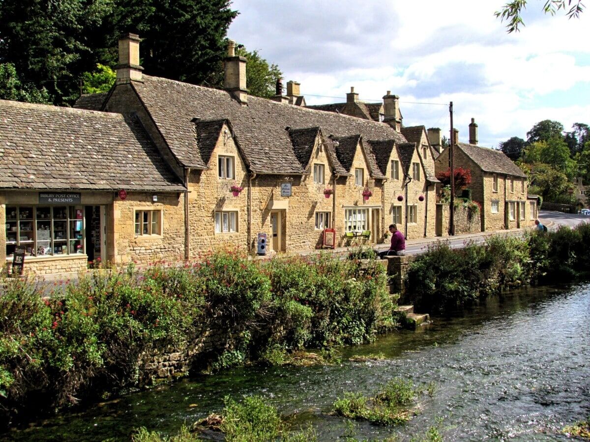 Row of charming stone cottages with sloped roofs along a riverbank, lush greenery in the foreground, and a person sitting by the water in a scenic village setting under a partly cloudy sky.