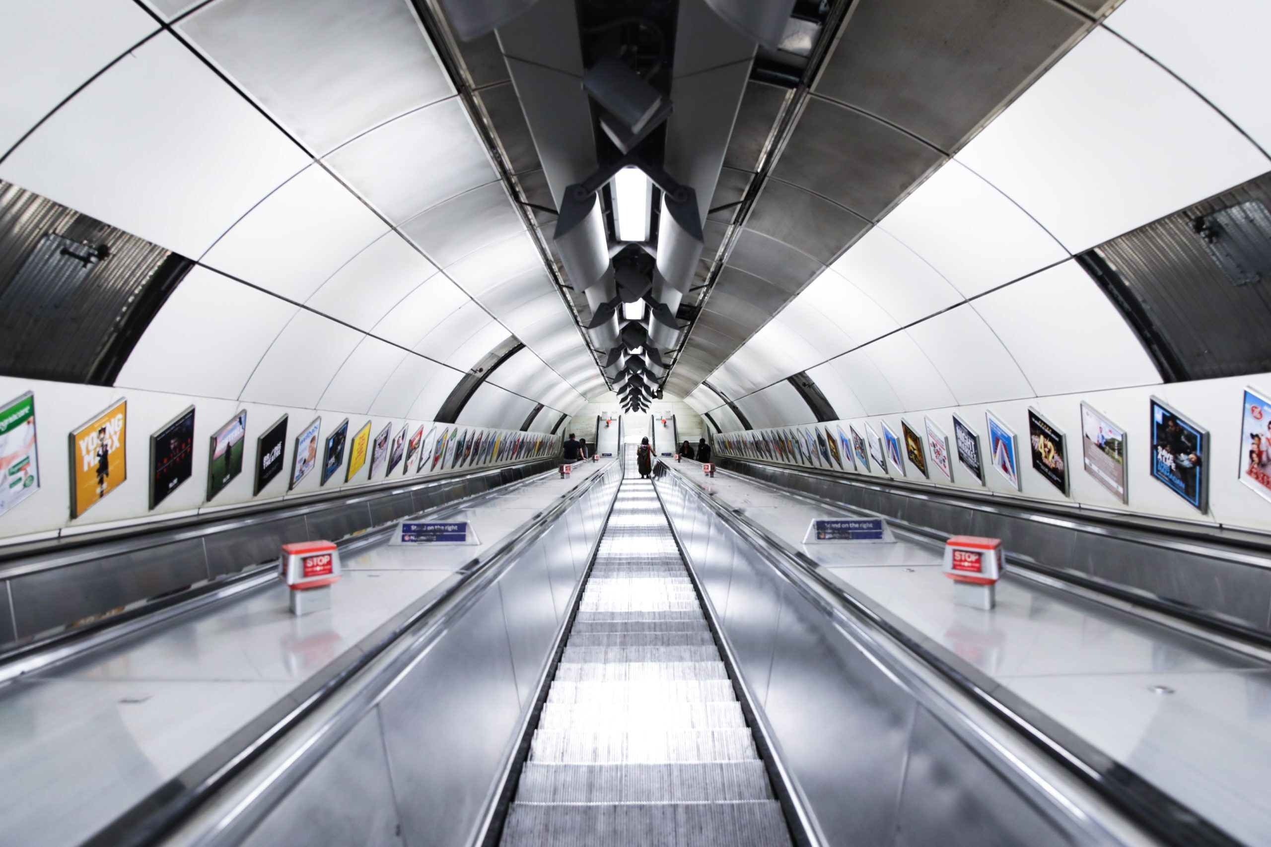 A wide, empty escalator tunnel in an underground subway station, with bright lighting, a curved ceiling, and colorful advertising posters lining both walls. A few people are visible in the distance at the far end.