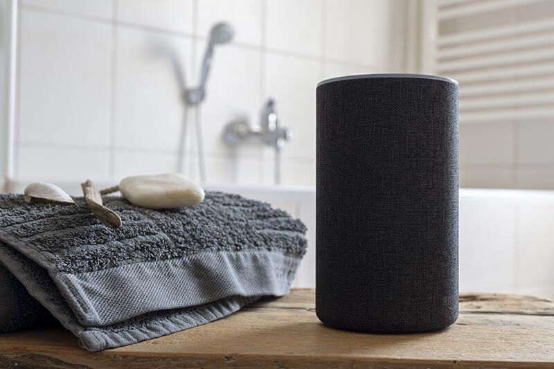 A smart speaker sits on a wooden surface next to folded gray towels, a toothbrush, and soap, with a bathtub and showerhead visible in the tiled bathroom background.