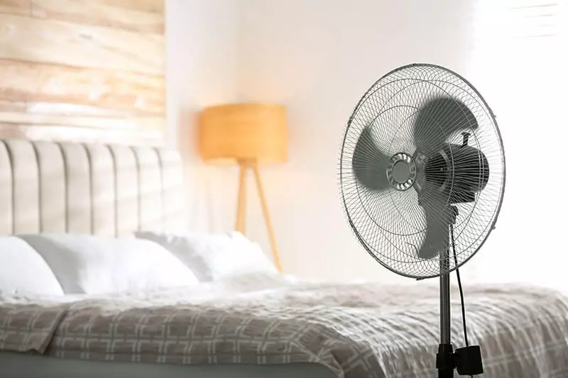 A standing electric fan is placed beside a bed with white bedding in a softly lit bedroom, featuring a wooden headboard and a floor lamp with a beige shade in the background.