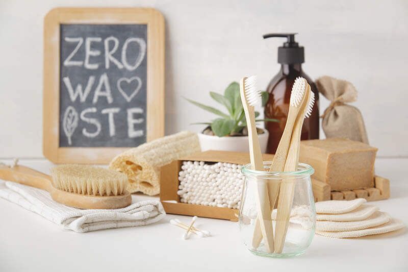 A collection of zero-waste bathroom essentials, including bamboo toothbrushes in a glass jar, a bar soap, a wooden brush, reusable cotton pads, and a chalkboard sign reading ZERO WASTE in the background.