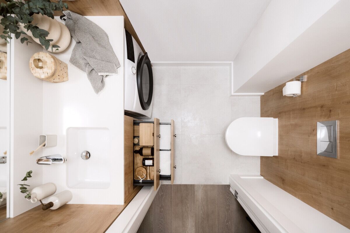 Modern bathroom with wood and white accents, seen from above. Features a sink with a mirror, a washing machine, open drawers with toiletries, a toilet, toilet paper holder, and a plant in a vase on the counter.