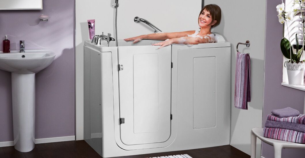 A smiling woman relaxes in a walk-in bathtub with white doors, surrounded by a modern bathroom featuring a sink, towels, and flowers on the windowsill.