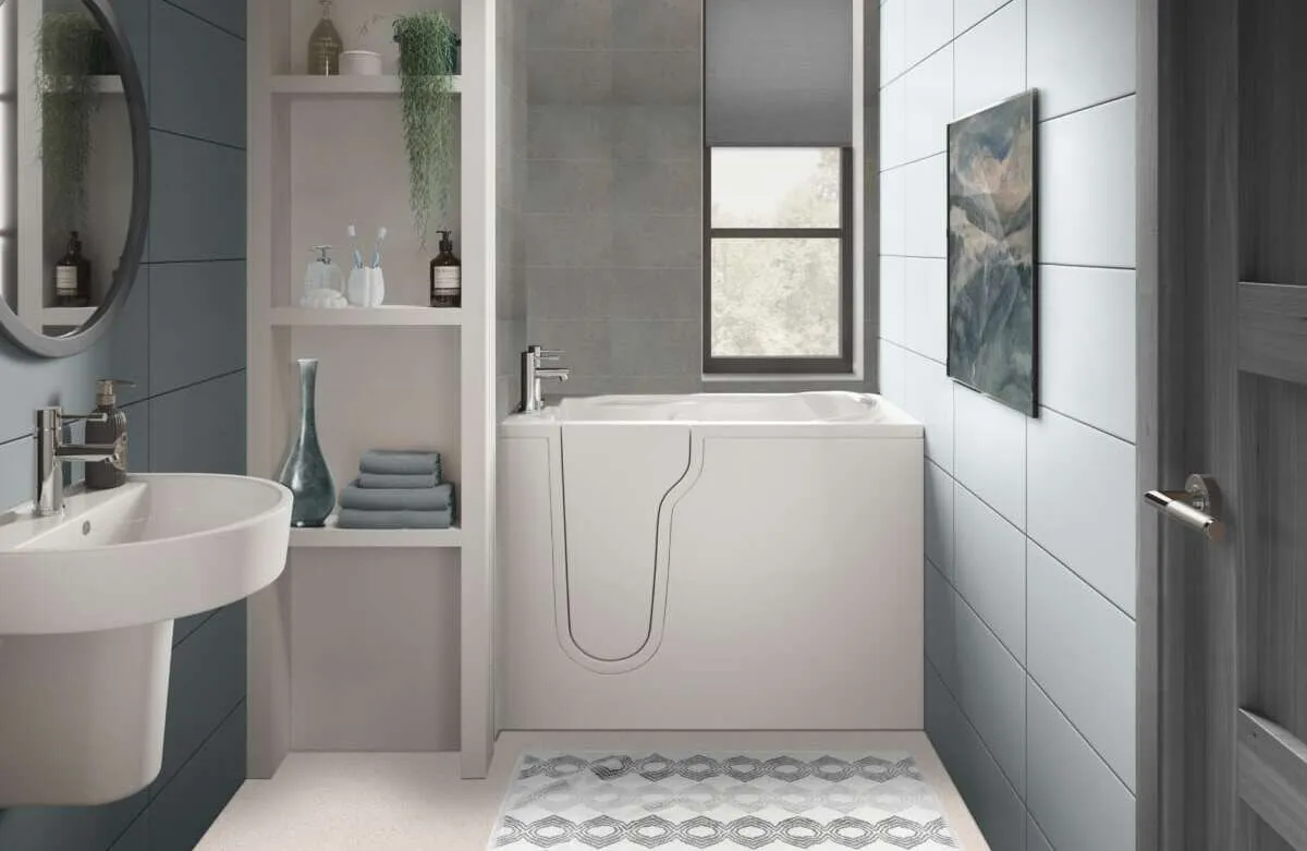 Modern bathroom with a walk-in bathtub, wall-mounted sink, open shelves with towels and toiletries, a round mirror, a window, and blue-gray tiled walls. A gray patterned rug is on the floor.