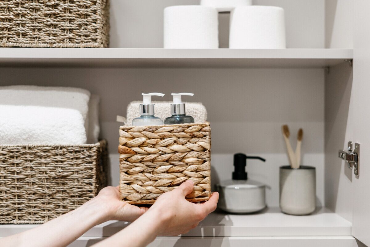 Hands placing a woven basket holding two pump bottles on a bathroom shelf, with rolled towels, toilet paper, a soap dispenser, and toothbrushes visible in the background.