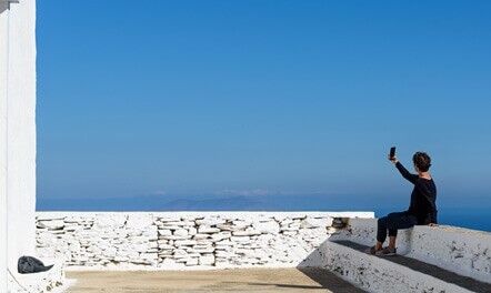 A person sits on white steps by a white stone wall, holding up a phone to take a selfie against a clear blue sky and distant sea.