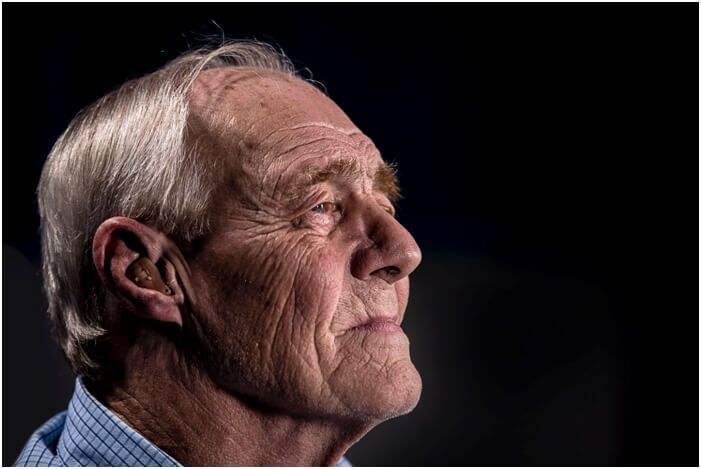 An elderly man with gray hair and deep wrinkles gazes thoughtfully into the distance. He wears a hearing aid in his ear and a blue shirt, with a dark background behind him.