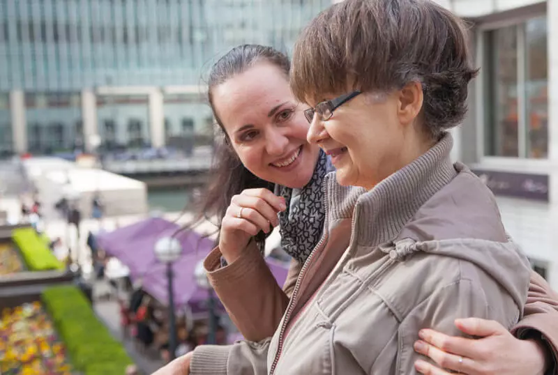 Two women stand outdoors, smiling and embracing. One woman wears glasses and a light jacket, while the other stands behind her, smiling warmly. An urban setting with buildings and purple awnings is visible in the background.