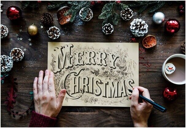 A person draws festive Merry Christmas lettering on paper, surrounded by pinecones, ornaments, and a cup of hot chocolate with a snowman marshmallow on a wooden table.