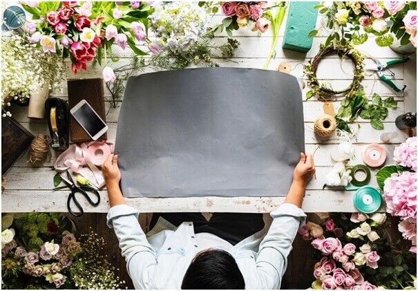 A person sits at a wooden table covered with flowers, ribbons, and craft supplies, holding a large blank sheet of black paper in the center, ready for a floral or creative project.