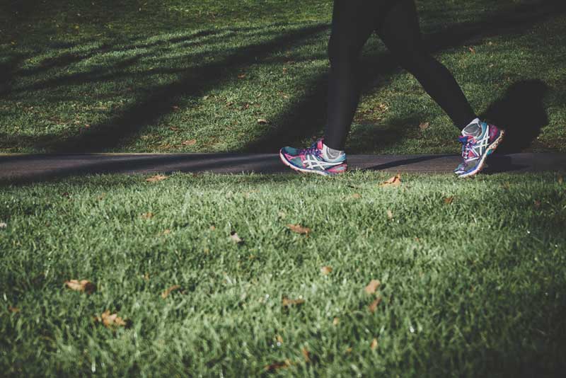 Person wearing black leggings and colorful running shoes walking on a paved path through a grassy area, with sunlight and shadows on the grass.
