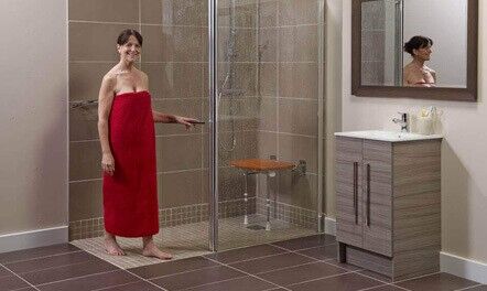 A woman wearing a red towel stands in a modern bathroom next to a walk-in shower with a fold-down wooden seat. A sink with a mirror is on the right. The bathroom has brown tile flooring and neutral-colored walls.