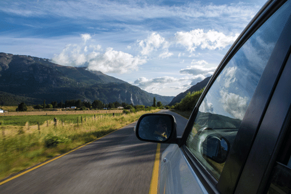 A car drives along a rural road with mountains, green fields, and clouds visible in the distance. The cars side mirror and window reflect the scenic landscape.