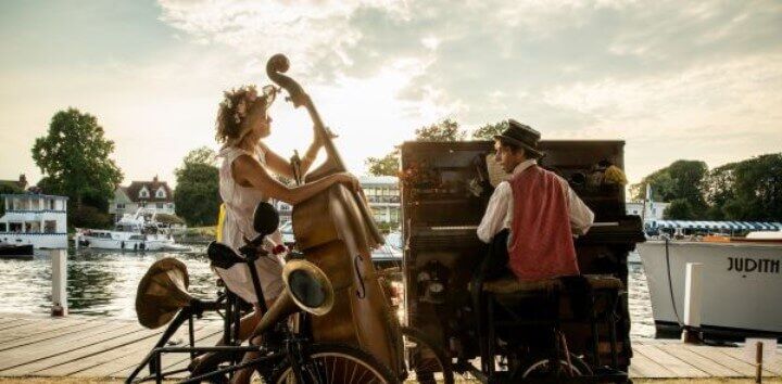 Two musicians perform outdoors by the water, one playing a double bass while seated on a bicycle, the other playing an upright piano. Boats and houses are visible in the background under a partly cloudy sky.