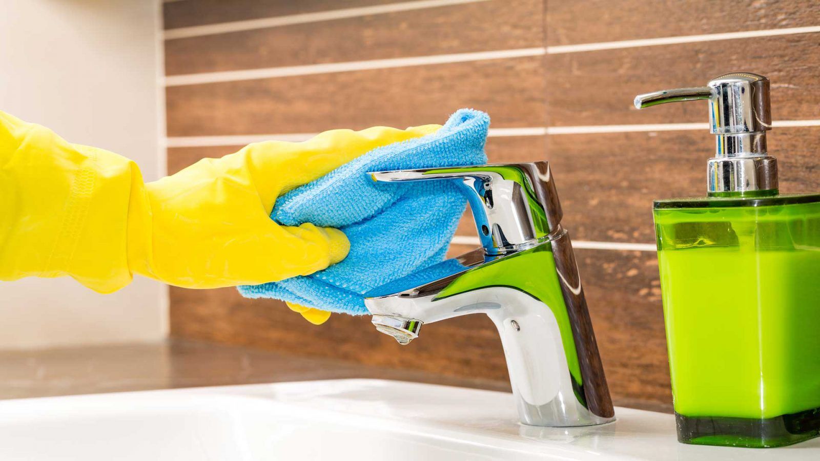 A person wearing yellow rubber gloves cleans a shiny bathroom tap with a blue cloth. A green soap dispenser is placed next to the tap on a white sink. The background features brown tiled walls.