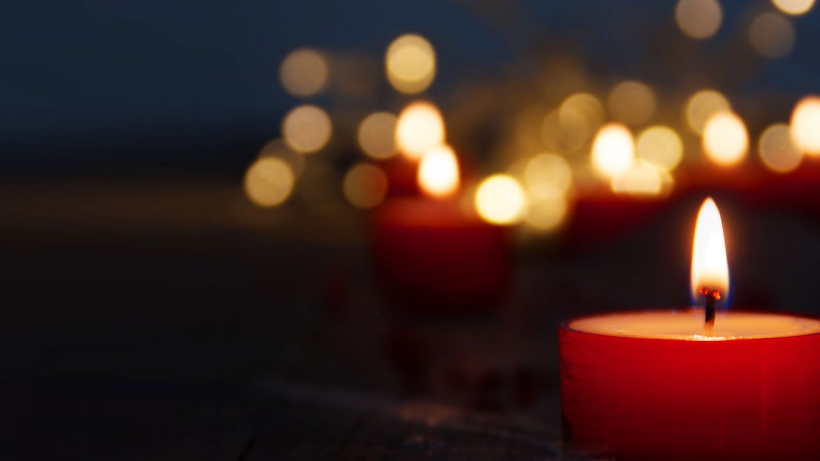 A close-up of a small red tealight candle burning with a bright flame, surrounded by several other out-of-focus candles and warm, glowing bokeh lights in the dark background.