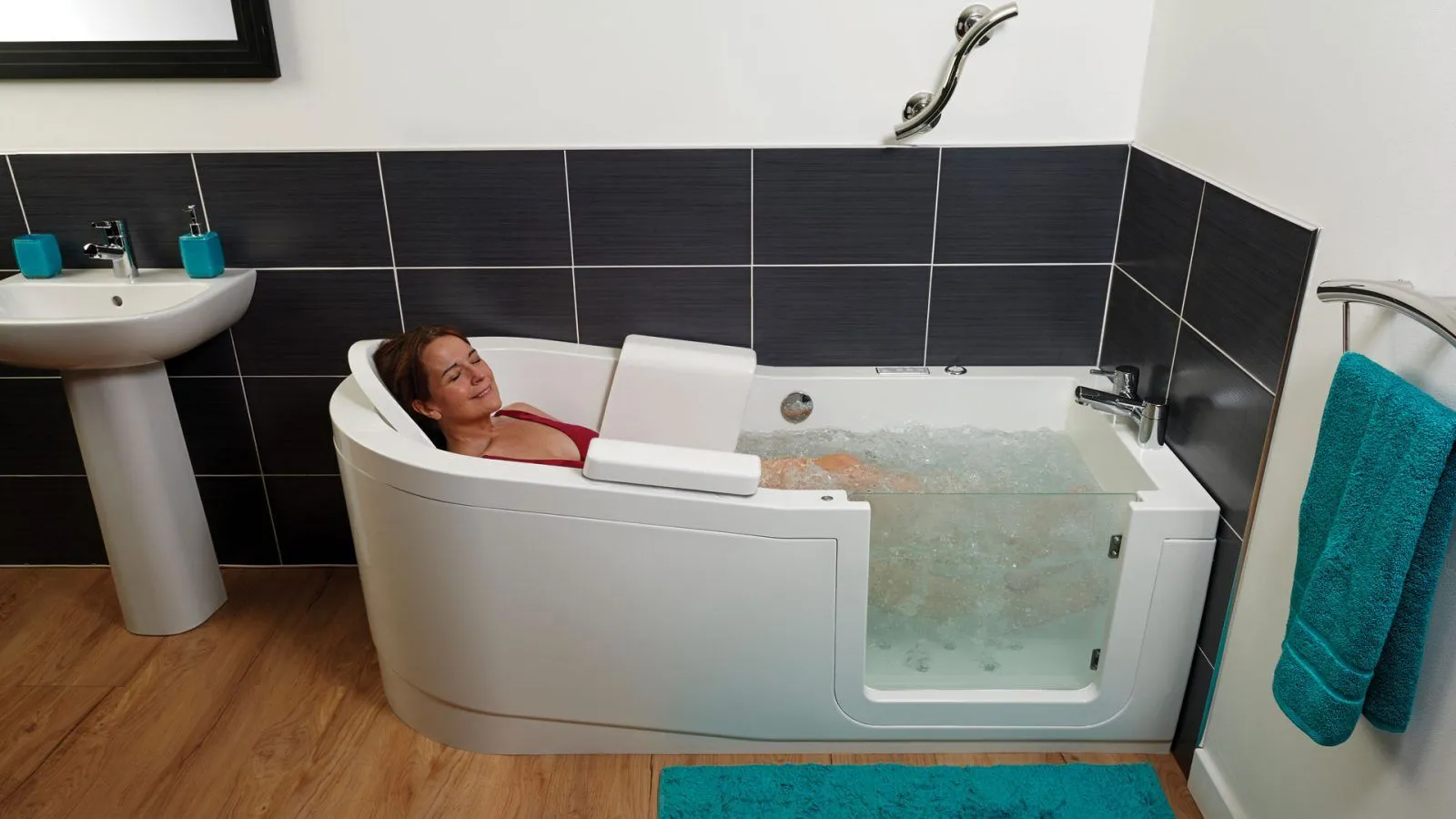 A woman relaxes in a modern walk-in bathtub with a glass door, surrounded by black tiles, a sink, and turquoise towels. The water in the tub is bubbly.