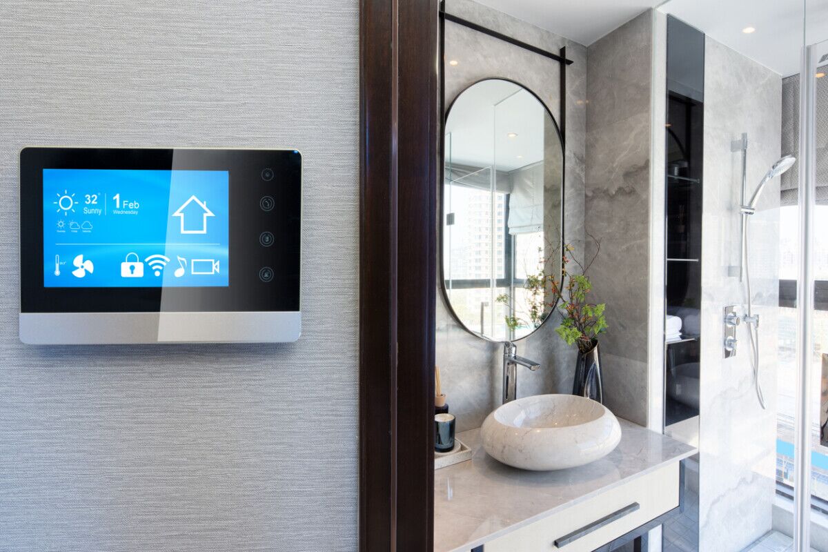 A smart thermostat displays weather and home info on a wall outside a modern bathroom with a round mirror, sink, and glass shower enclosure. The bathroom features light gray walls and contemporary decor.