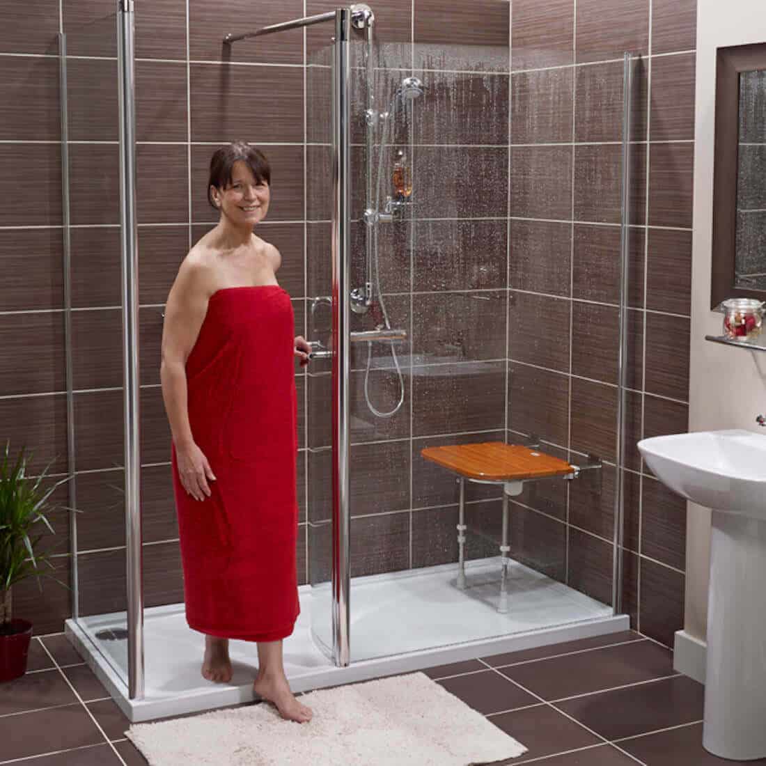 A woman wearing a red towel stands at the entrance of a modern walk-in shower with a glass door and a wooden shower seat. The bathroom has brown tiled walls, a plant, and a white sink.