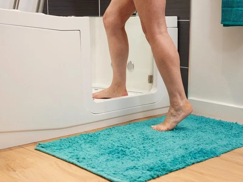 A person steps into a walk-in bathtub, with one foot inside and one foot outside on a turquoise bath mat, in a bathroom with wood flooring and tiled walls.