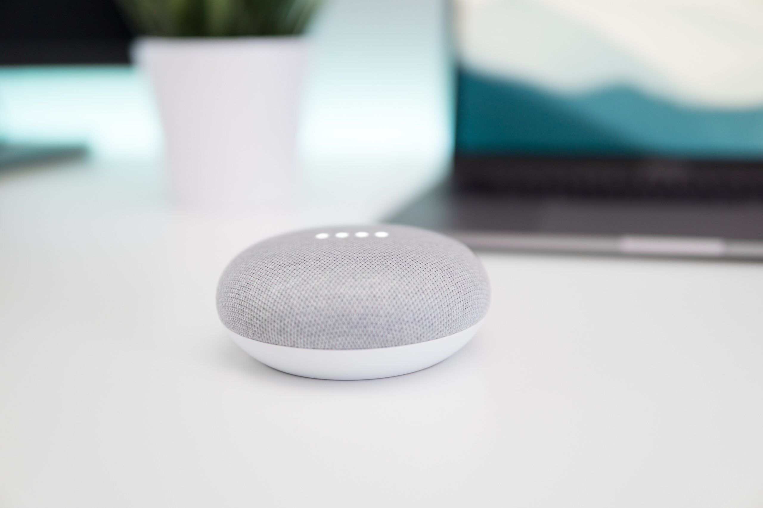 A gray smart speaker with four white LED lights sits on a white desk near a laptop and a potted plant in a blurred background.