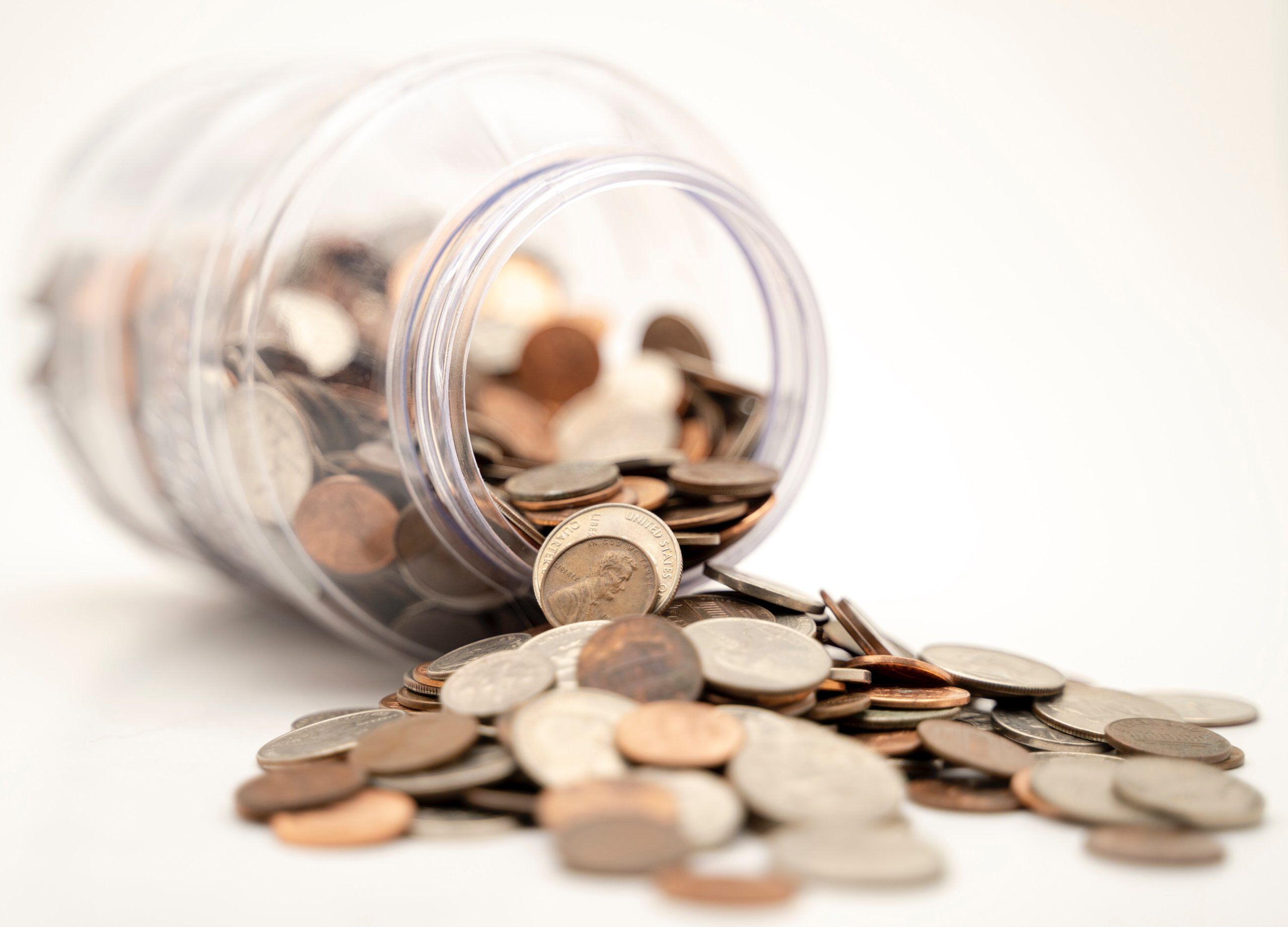 A clear jar tipped over on its side spills assorted coins onto a white surface.