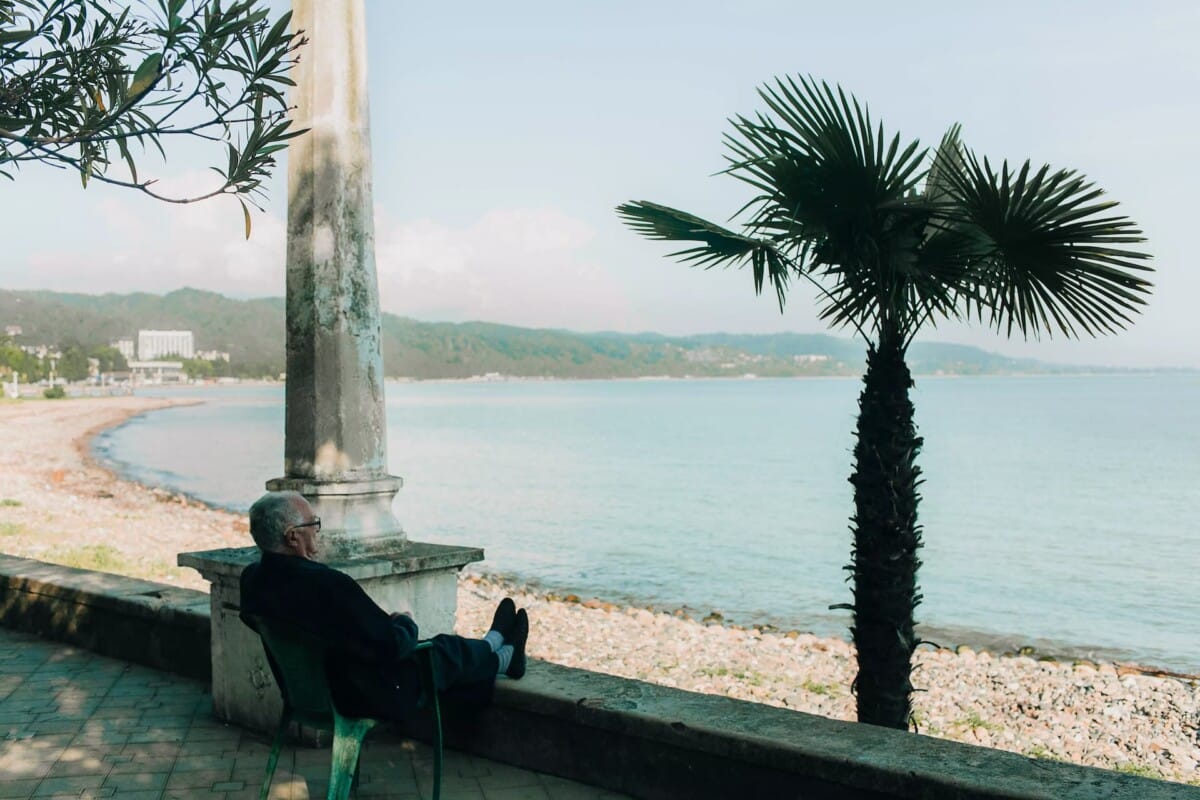 An elderly man sits on a chair by a stone railing, relaxing and gazing out at a calm, rocky coastline lined with palm trees on a partly cloudy day.